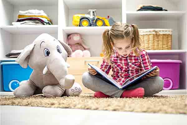 little girl sitting on the carpet and reading a book for her stuffed elephant