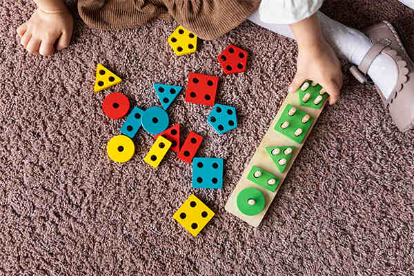 A child playing with shape-sorting educational toys on a carpeted floor, surrounded by vibrant geometric pieces