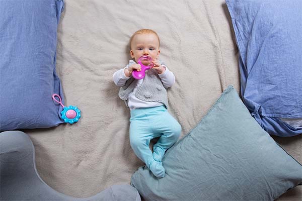 Sweet red haired baby lying on blanket among pillows and playing with teething toys 