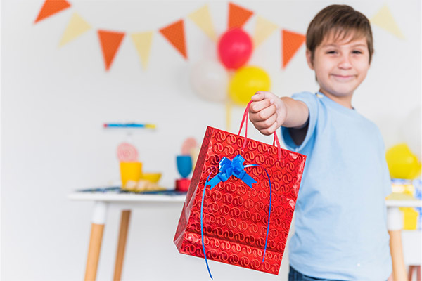 A smiling boy giving a red birthday gift bag to someone
